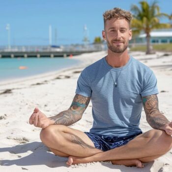 Man with tattoos meditating on a sunny beach with clear skies and palm trees in the background