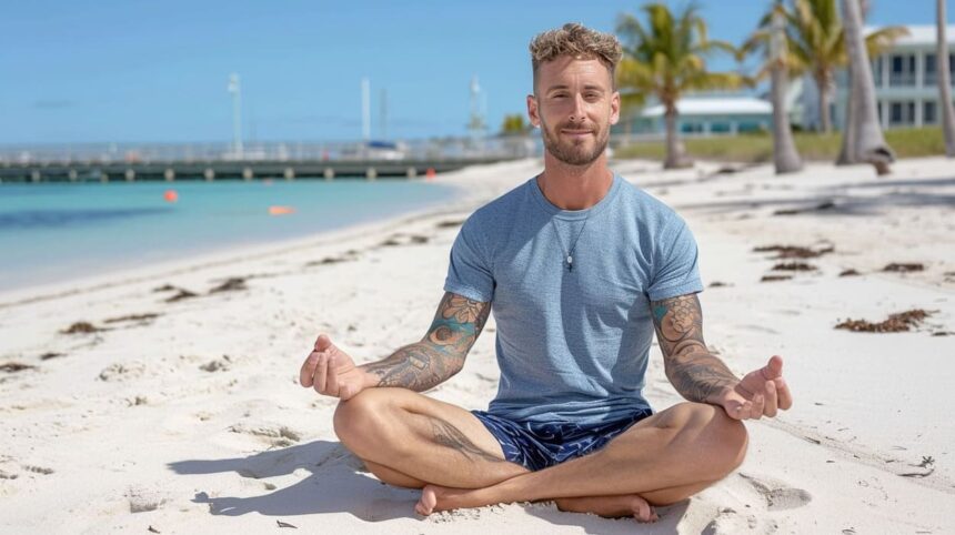 Man with tattoos meditating on a sunny beach with clear skies and palm trees in the background