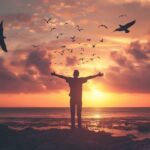 Person with outstretched arms standing on the beach at sunset with birds flying in the sky.