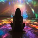 Woman sitting on floor watching colorful light projection in dark room.