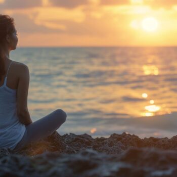 Woman sitting peacefully on the beach at sunset looking at the ocean.