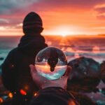 Person holding crystal ball with sunset reflected over rocky coastline.
